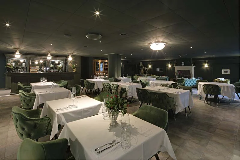 Foto eines Restaurants mit dunklen Wänden, grünen Samtsesseln und Tischen mit weissen Decken